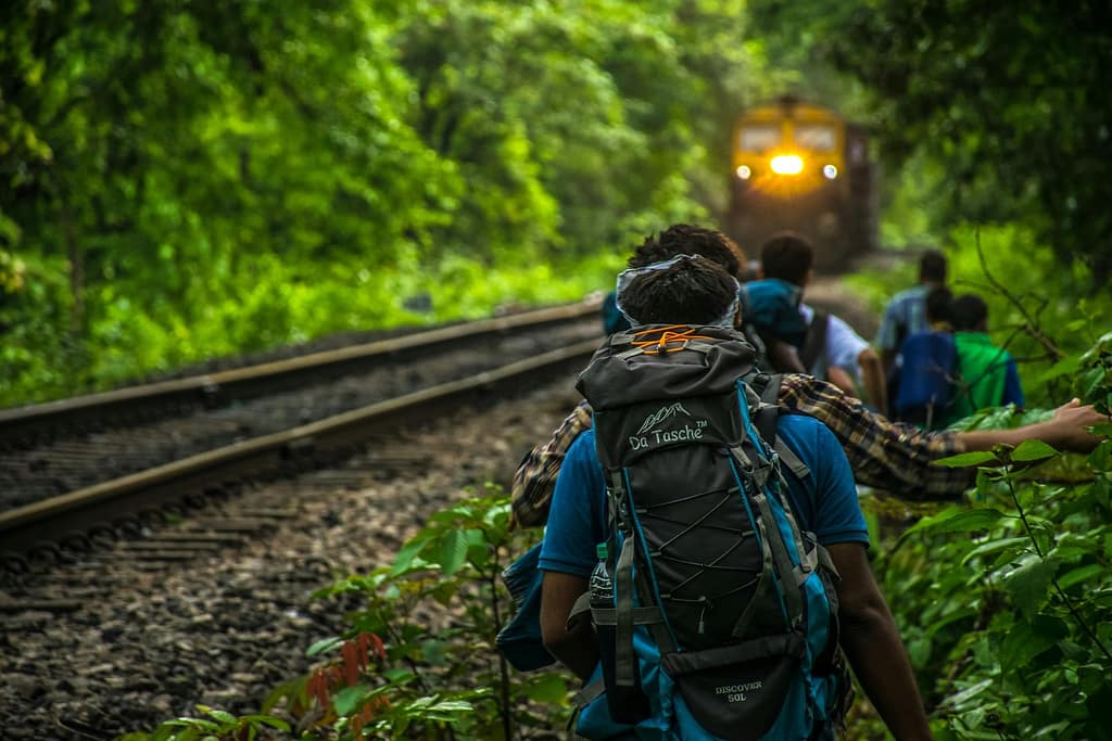 Group of backpackers walking along railway tracks through lush forest with approaching train.